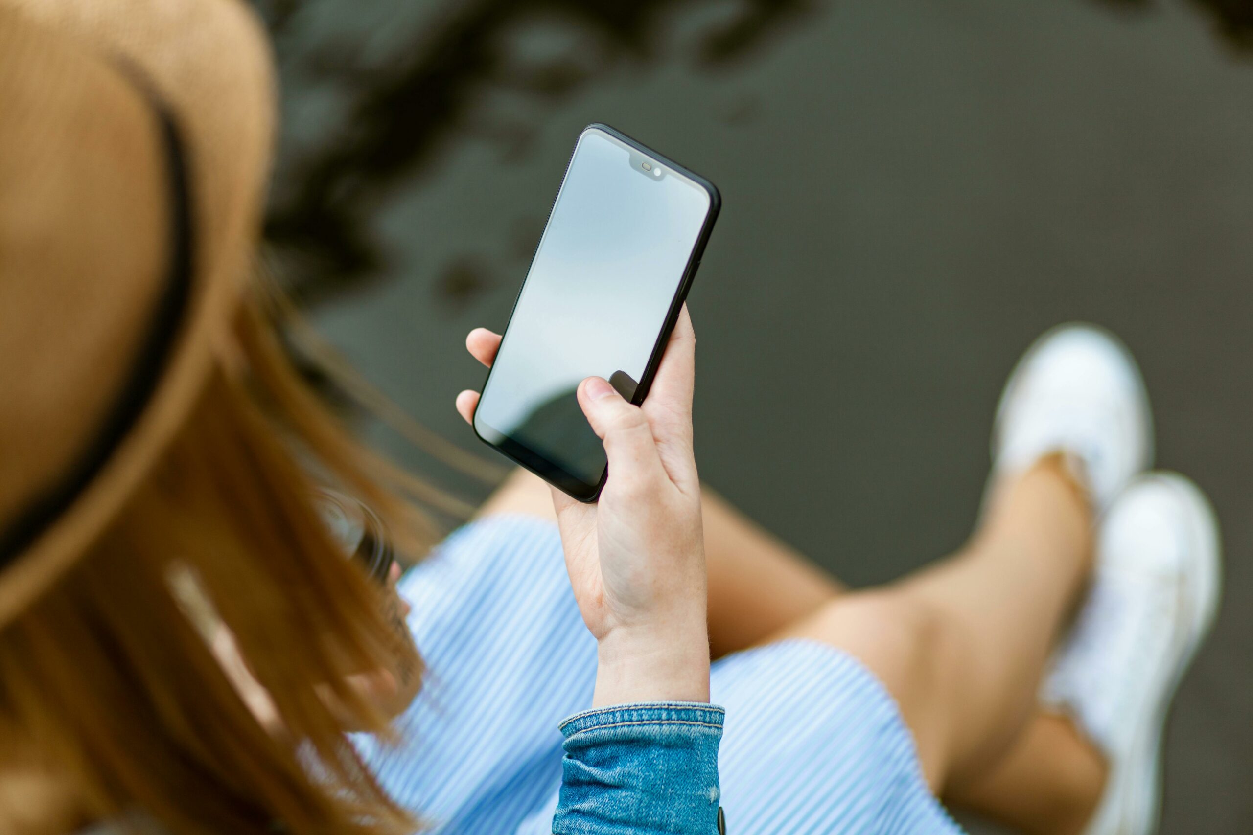 A young woman holding a smartphone while sitting outdoors in summer, capturing a moment of relaxation and technology use.